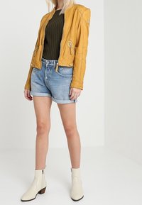 Gul läderjacka, mörkgrön ribbad tröja, denimshorts och vita ankellånga stövlar med brun klack. Slät textur och kroppsnära design.