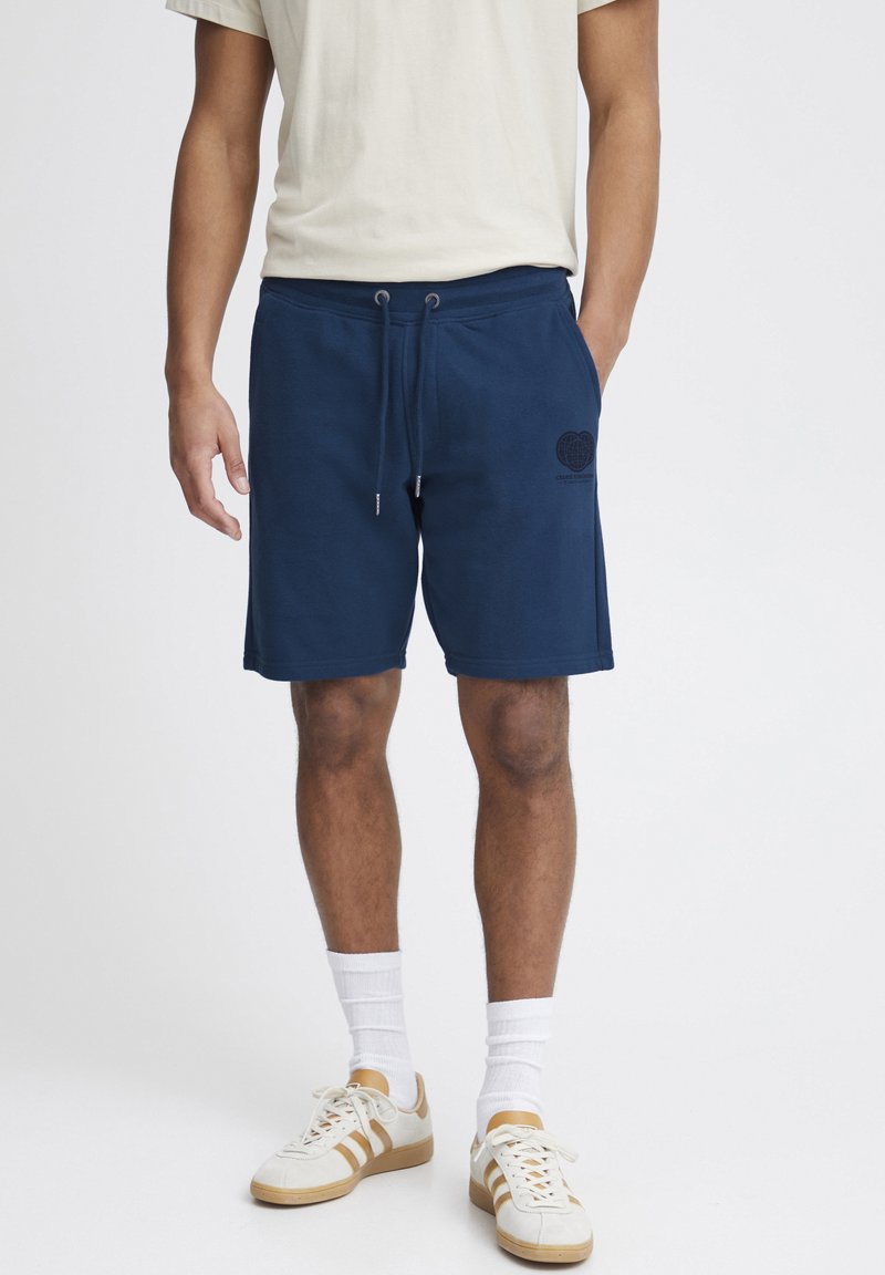 Shorts en coton bleu marine avec une taille ajustable à cordon, arborant un logo cœur texturé sur la jambe gauche. Baskets blanches et chaussettes.