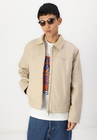 Chaqueta beige con cuello y cierre de cremallera, que presenta una tela texturizada y sutilezas en las costuras. Se lleva sobre una camiseta blanca con gráfico, combinada con jeans.