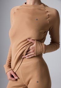Person wearing beige long-sleeve top and matching pants, with one hand lifting the top at the waist, showing fitted clothing details.