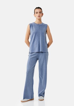 Frau steht mit Händen hinter dem Rücken, trägt ärmelloses blaues gestricktes Oberteil und passende weite Hosen mit weißen Sneakern vor einfarbigem Hintergrund.