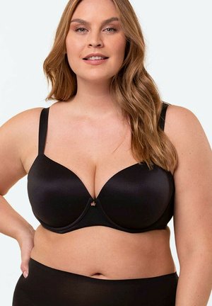 Uśmiechnięta kobieta w rozmiarze plus size z długimi, falowanymi włosami, ubrana w czarny biustonosz i czarne majtki na jasnym tle.