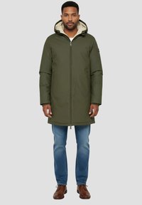 Chaqueta verde oliva con capucha, cierre de cremallera, bolsillos laterales y forro acolchado. Combinada con vaqueros azules y zapatos marrones.