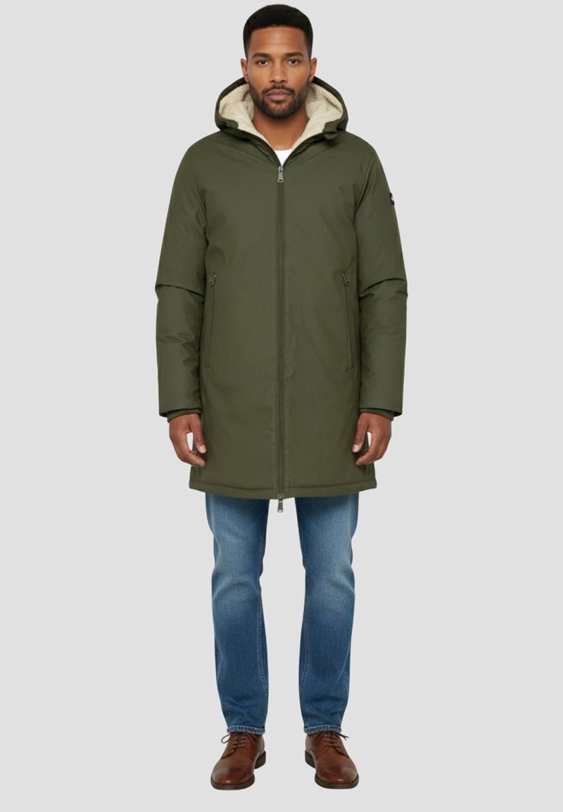 Chaqueta verde oliva con capucha, cierre de cremallera, bolsillos laterales y forro acolchado. Combinada con vaqueros azules y zapatos marrones.