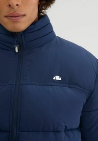 Chaqueta acolchada azul marino con cuello alto, costuras horizontales y un pequeño logo blanco en la parte superior izquierda. Tejido suave y aislante.