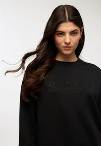 Felpa nera con il testo "SELFLOVE Club" in un motivo ricamato coordinato; presenta un collo rotondo e maniche lunghe. Tessuto con una texture liscia.