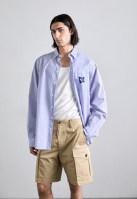 Camisa de botones a rayas en azul y blanco con un logo en el pecho, combinada con pantalones cortos de carga beige con bolsillos laterales y un ajuste relajado.