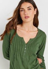 Groene langoesche mouwtop met een ronde halslijn, knoopsluiting en verticale plooien aan de voorkant, gemaakt van zachte, getextureerde stof.