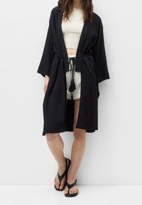 Kimono negro texturizado con mangas anchas, atado a la cintura con un cordón y detalle de borlas. Usado sobre un top corto ligero y pantalones cortos.