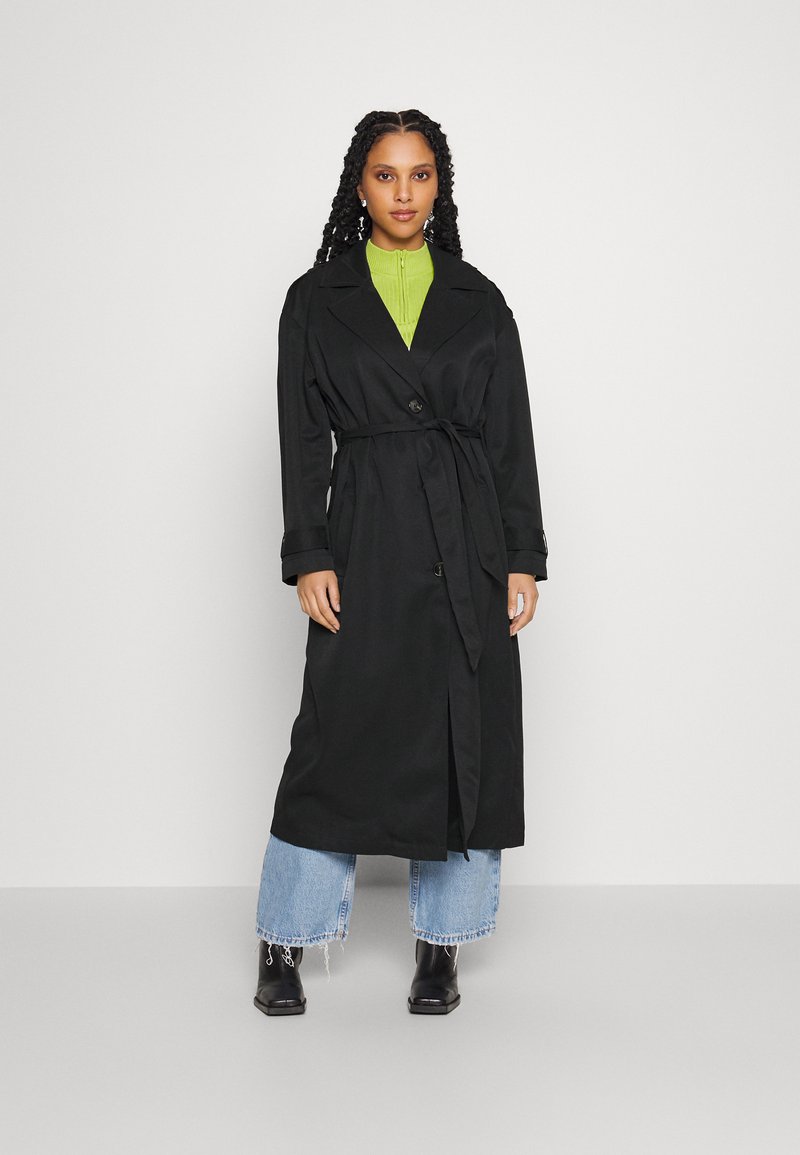 Svart trench coat med belte, dobbeltspent design og lange ermer. Båret over en grønn topp og parret med blå cropped jeans.