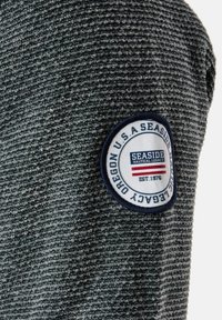 Texturerad grå stickad tröja med en vit cirkulär lapp som har "Seaside" och "Nautical Legacy"-designs, marinblå kant på lappen.