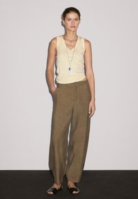 Il modello sta in piedi con sicurezza indossando una canotta gialla chiara e pantaloni verde oliva, con una lunga collana e sandali neri, su uno sfondo grigio.