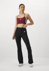 Burgunderfärgad sport-bh med tunna remmar och vita tre-rands detaljer; svarta utsvängda leggings med en liten vit logotyp på höften.