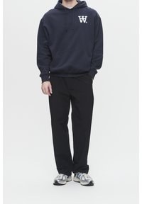 Marineblauer Kapuzenpullover mit weißem Logo, kombiniert mit schwarzer Hose und grauen Sneakern. Der Kapuzenpullover hat eine Kängurutasche und eine Kapuze mit Kordelzug.