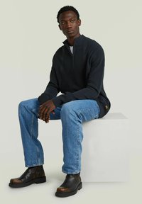 Man die op een wit blok zit, gekleed in een zwart half-zip sweater, lichtblauwe jeans en zwarte laarzen, en iets opzij kijkt.