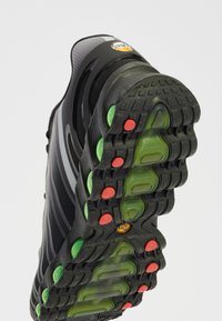 Sapatilha desportiva preta com um upper texturizado, apresentando um logótipo proeminente e uma sola de borracha grossa com pontos de tração verde e vermelho.