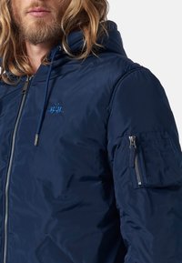 Marinblå vadderad jacka med huva, har dragkedjefront, sidfickor och en ärmficka med dragkedja. Slät, glänsande textur med broderad logotyp.