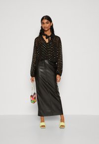 Schwarze, transparente Bluse mit bunten Punkten und gebundenem Kragen; figurbetonender schwarzer Leder-Maxi-Rock; knallgrüne Absatzsandalen; handgefertigte mehrfarbige Handtasche.