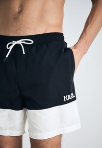 Svart og hvit svømmebukse med elastisk midje, snøring og logo-detalj. Laget av lett materiale, med kontrastfargeblokkerte design.