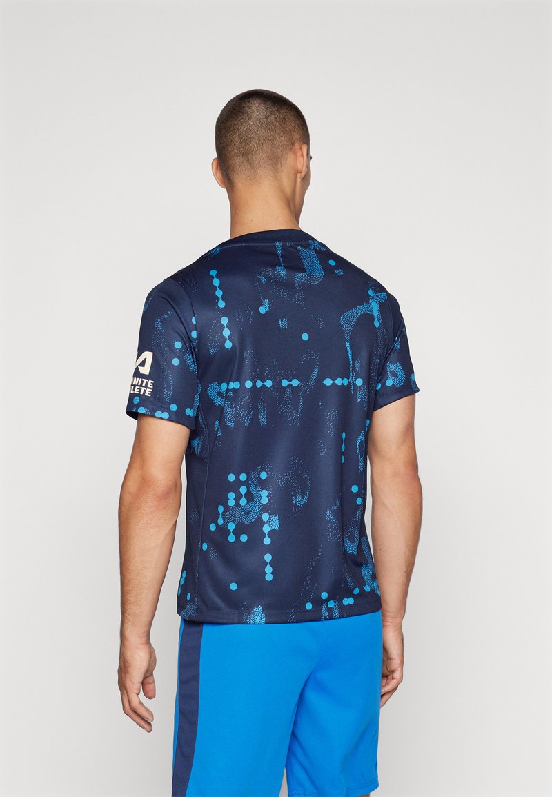 Maglietta sportiva navy con motivi a pois blu, maniche corte e collo tondo. Presenta un logo sul braccio sinistro. Indossata con pantaloni corti blu.