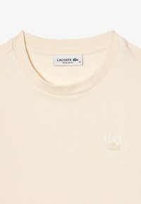 Camiseta color crema hecha de tela suave, con cuello redondo y logotipo en relieves en el pecho. Diseño de ajuste regular.