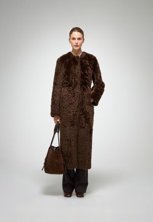 Pruun pikk mantel, mis on valmistatud pehmest tekstuurse materjaliga, millel on ümmargune krae ja küljetaskud. Sobitatud tumedate pükste ja pruuni käekotiga.