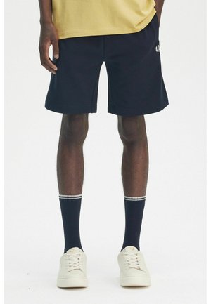 Pantaloni corti neri in misto cotone, con vita elastica e tasche laterali, abbinati a sneakers bianche e calzini blu navy con strisce.