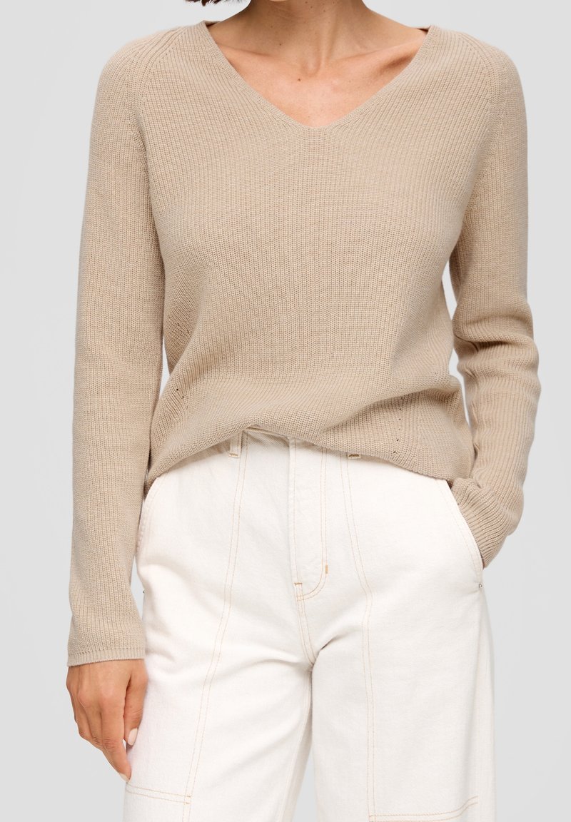 Pull beige tricoté avec un col en V, texture côtelée et manches longues, associé à un pantalon blanc taille haute avec des détails de couture visibles.