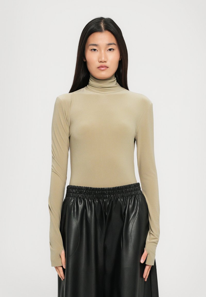 Donna con lunghi capelli lisci, che indossa un maglione beige a collo alto e una gonna in pelle nera a vita alta, in piedi davanti a uno sfondo semplice.