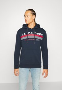 Sudadera navy con bolsillo frontal, con un diseño de rayas rojas y blancas y el texto "JACK & JONES". La tela parece suave y casual.