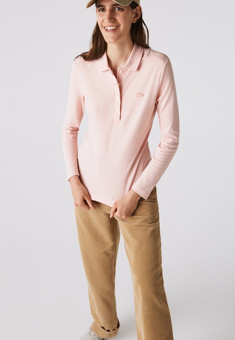 Frau lächelnd, trägt ein hellrosa langärmeliges Poloshirt, beige Cordhosen, beige Kappe und weiße Turnschuhe, steht vor einem einfarbigen Hintergrund.