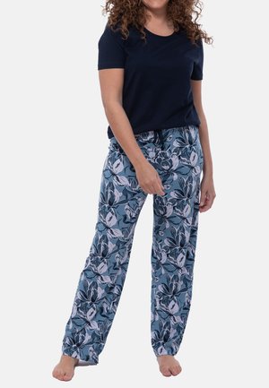Frau in einem navyblauen Kurzarmhemd und weit geschnittenen, blauen Blumenmusterhosen, die barfuß vor einem weißen Hintergrund steht.