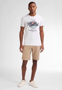 Maglietta bianca a maniche corte con un design grafico multicolore e testo. Indossata con pantaloncini color crema e scarpe da ginnastica di colore chiaro. Sfondo neutro.