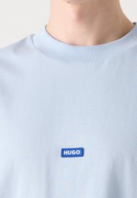Homme portant un t-shirt bleu clair à col rond avec un petit patch rectangulaire bleu marqué "HUGO" sur la poitrine.