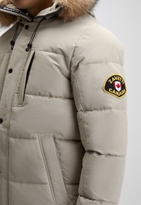 Veste matelassée beige avec une capuche en fourrure, présentant un logo sur la manche, des poches avant et une texture matelassée.