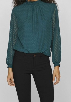 Vrouw draagt een turquoise doorschijnende blouse met lange mouwen met een chevronstructuur en geplooide kraag, gecombineerd met zwarte aansluitende broek.