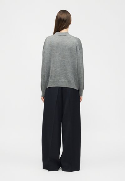 Άτομο με μακριά καστανά μαλλιά, που στέκεται με την πλάτη προς το μέρος μας, φοράει γκρι πουλόβερ με μακριά μανίκια και φαρδιά, μαύρα παντελόνια.