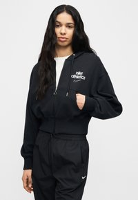 Νεαρή γυναίκα με μακριά σκούρα μαλλιά, φορώντας μαύρο φούτερ με φερμουάρ Nike Athletics και μαύρο παντελόνι φόρμας Nike, στέκεται μπροστά σε απλό φόντο.