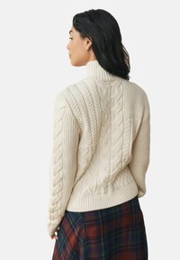 Cream cable-knit sweater with a high collar. Features ribbed and textured patterns, and a fitted hem. Paired with a plaid skirt.