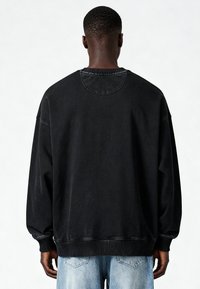 Man die met de rug naar de camera staat, gekleed in een losse zwarte trui en lichtblauwe jeans, tegen een effen witte achtergrond.