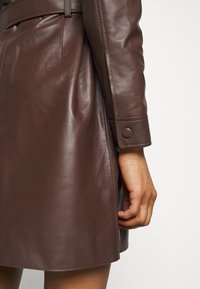 Brun læderjakke med en tætsiddende pasform. Har en sidesprække, knaplukning på ærmerne og en glat tekstur, der fremhæver materialets glans.