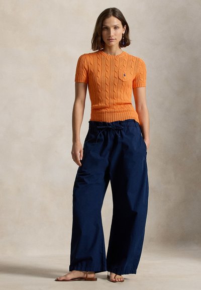 Γυναίκα που στέκεται ξυπόλητη φορώντας πορτοκαλί πλεκτό τοπ με κοντά μανίκια και σκούρο μπλε παντελόνι με φαρδιά πόδια, με τα χέρια στις τσέπες.