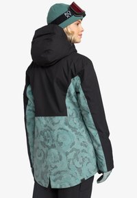 Schwarze und türkisfarbene Skijacke mit einem floralen Muster im unteren Bereich, ausgestattet mit Kapuze und Akzentdetails. Handschuhe sind ebenfalls sichtbar.