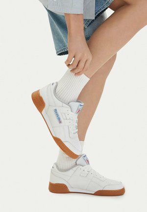 Osoba nosząca białe sneakersy Reebok z gumowymi podeszwami i białe prążkowane skarpetki, ubrana w krótkie dżinsowe spodenki i paskowaną koszulę.