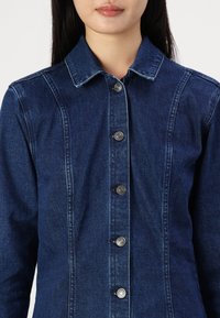 Camisa de mezclilla azul oscuro con cuello puntiagudo, cierre frontal de botones con herrajes plateados y detalles de costura vertical. Textura suave.