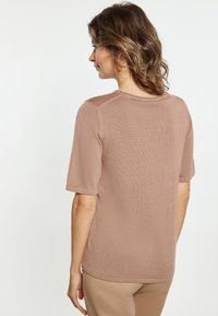 Korte mouw, ronde hals top in lichtbruin, met ribbelstructuur. Zachte stof met subtiele naden, gedragen met bijpassende beige broek, gezien van achteren.