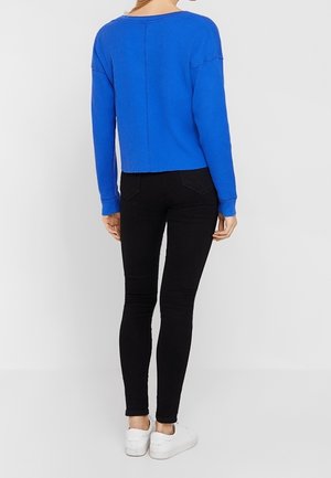 Woman standing with back to camera wearing a bright blue long-sleeve sweater, black skinny jeans, and white sneakers on a plain background.