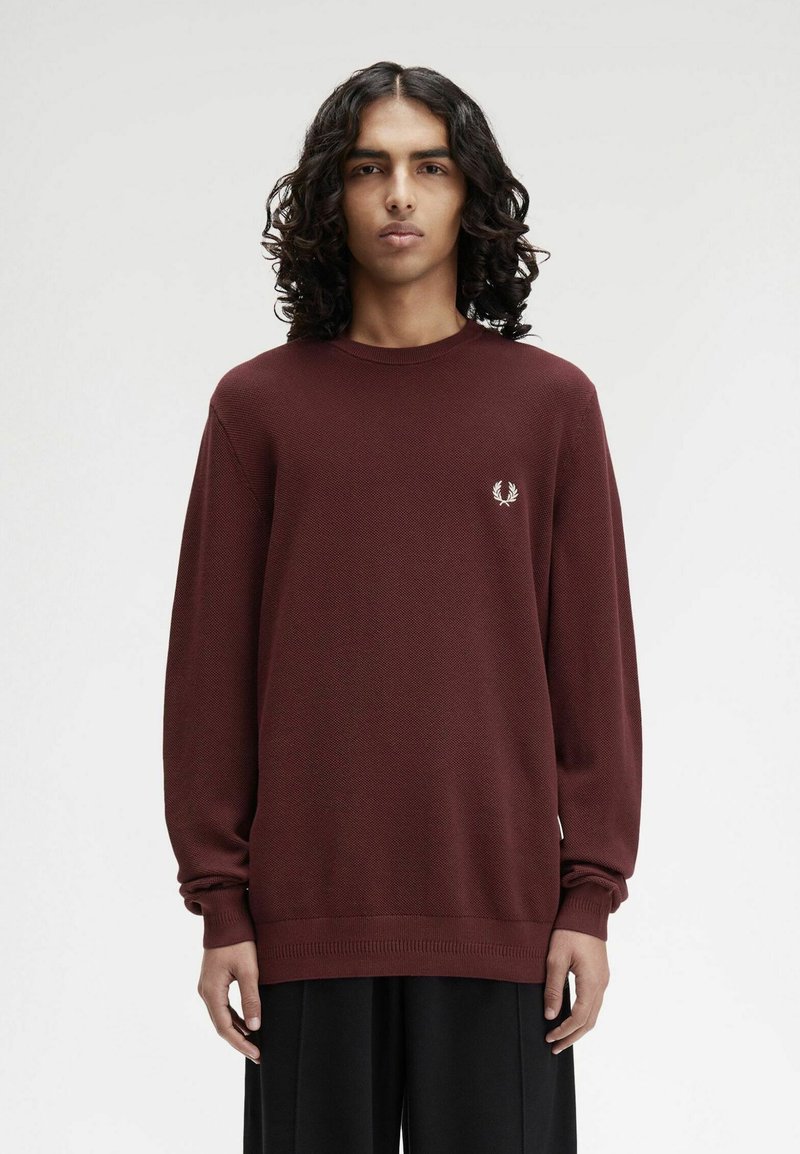 Persona con capelli ricci e lunghi che indossa un maglione bordeaux a maniche lunghe con un piccolo logo di alloro bianco, in piedi contro uno sfondo chiaro e neutro.