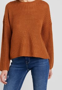 Maglione a maglia color ruggine con texture a coste, scollatura rotonda e spalle scese, abbinato a jeans in denim blu.