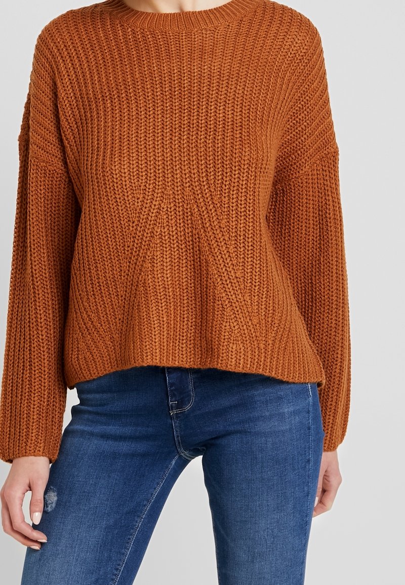 Maglione a maglia color ruggine con texture a coste, scollatura rotonda e spalle scese, abbinato a jeans in denim blu.